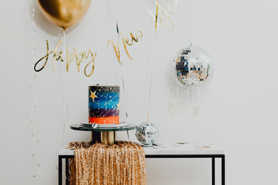Party Decorations - New Years sign, balloons, cake, disco ball - all placed in front of the white wall.
