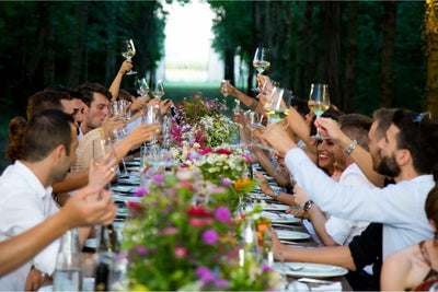 Group of friends toasting at a long outdoor dinner table adorned with flowers.